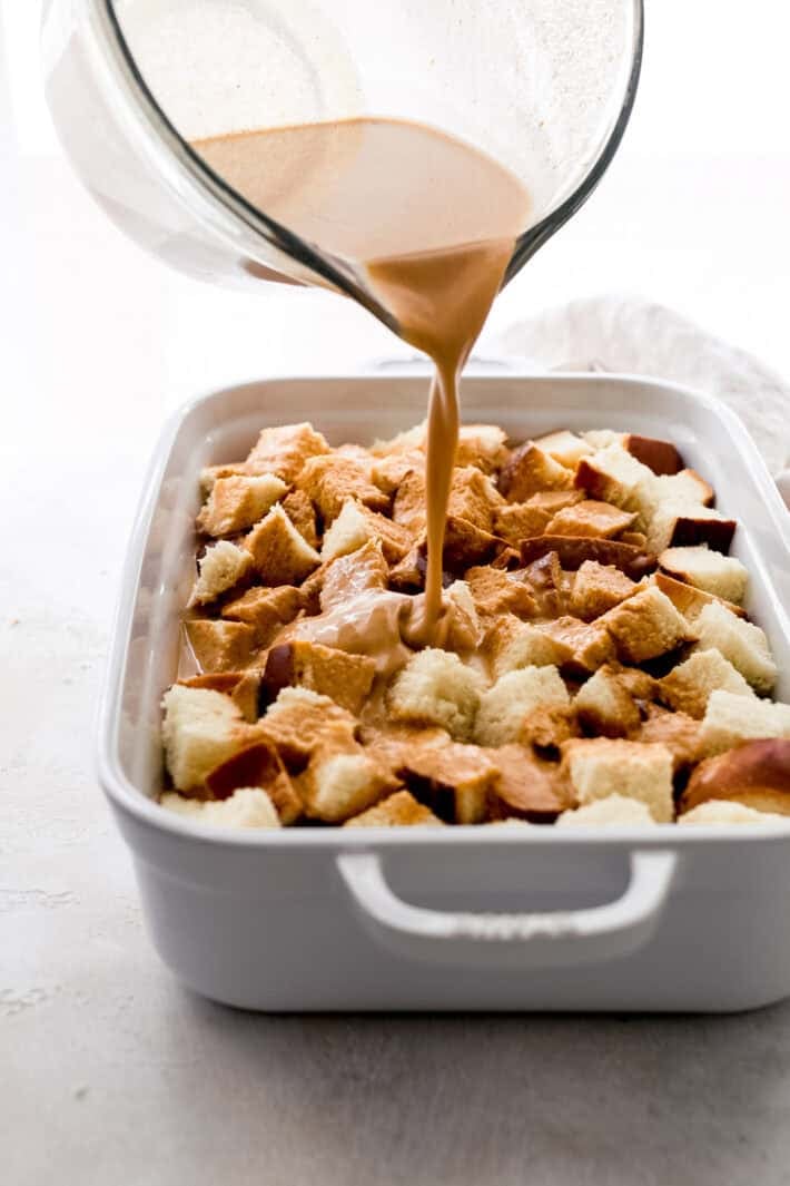pouring pumpkin custard mixture over bread cubes in white baking dish