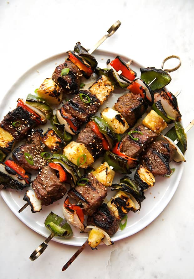 Sliced beef bulgogi on metal skewers showing deeply caramelized edges and visible char from the grill, glistening with the sweet-savory marinade, garnished with sesame seeds and sliced green onions on a black slate plate.