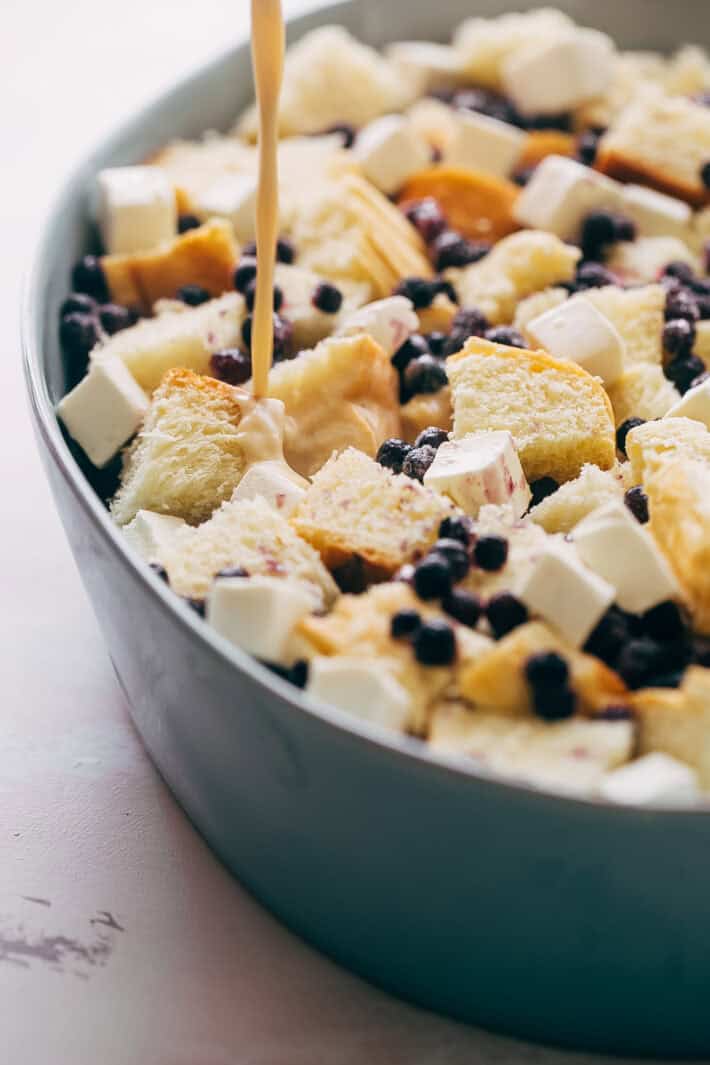 pouring egg batter into bowl with bread, cream cheese, and frozen blueberries