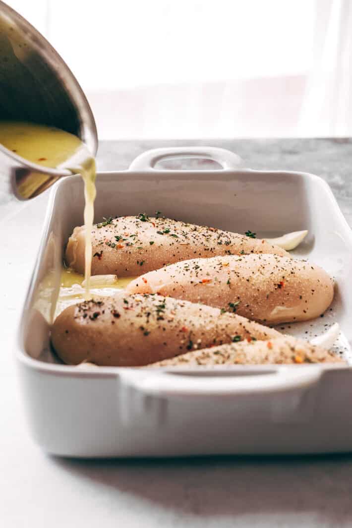 pouring prepared butter sauce on raw seasoned chicken in white baking dish